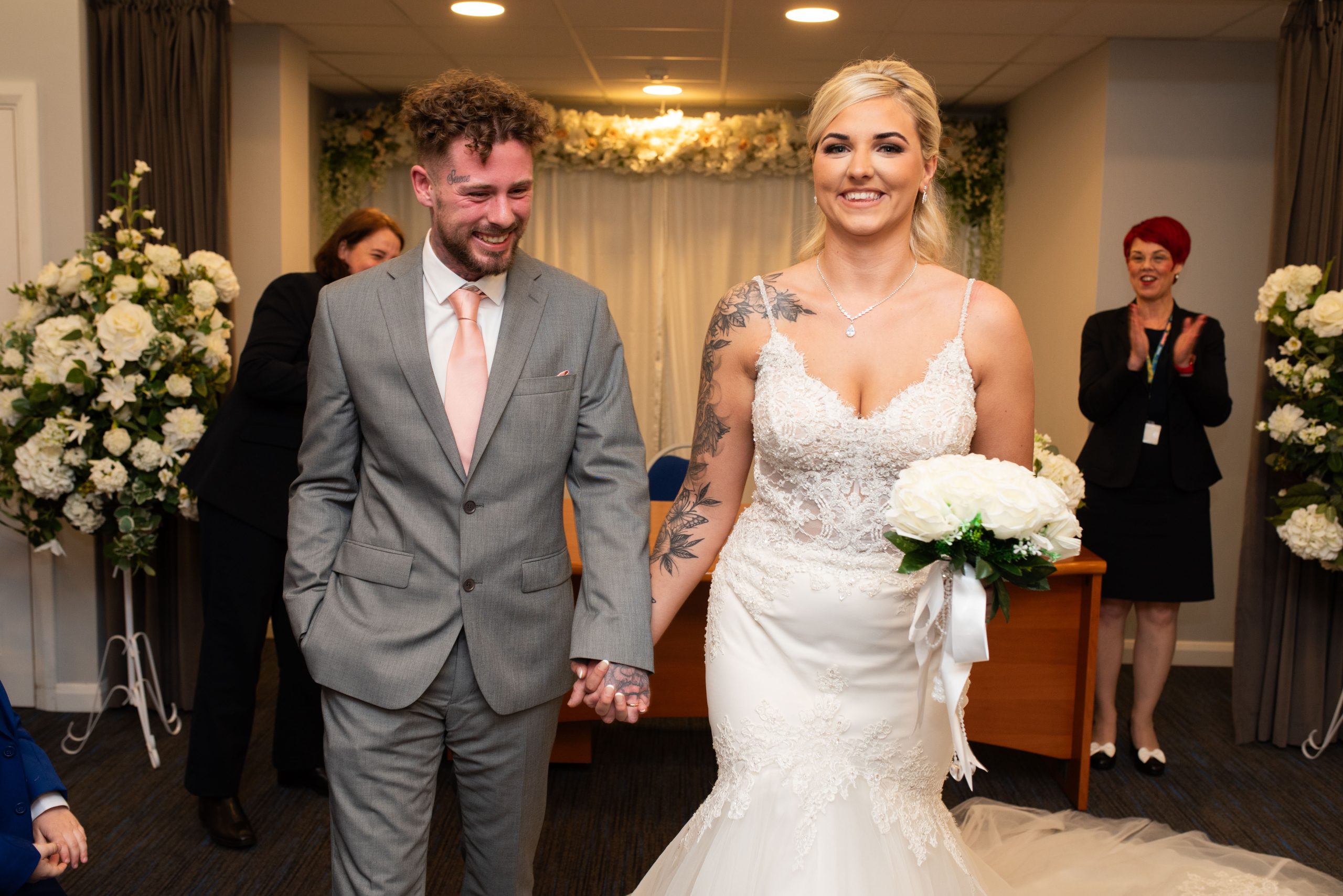 Newlyweds hold hands and smile at their wedding ceremony, with a registrar in the background.