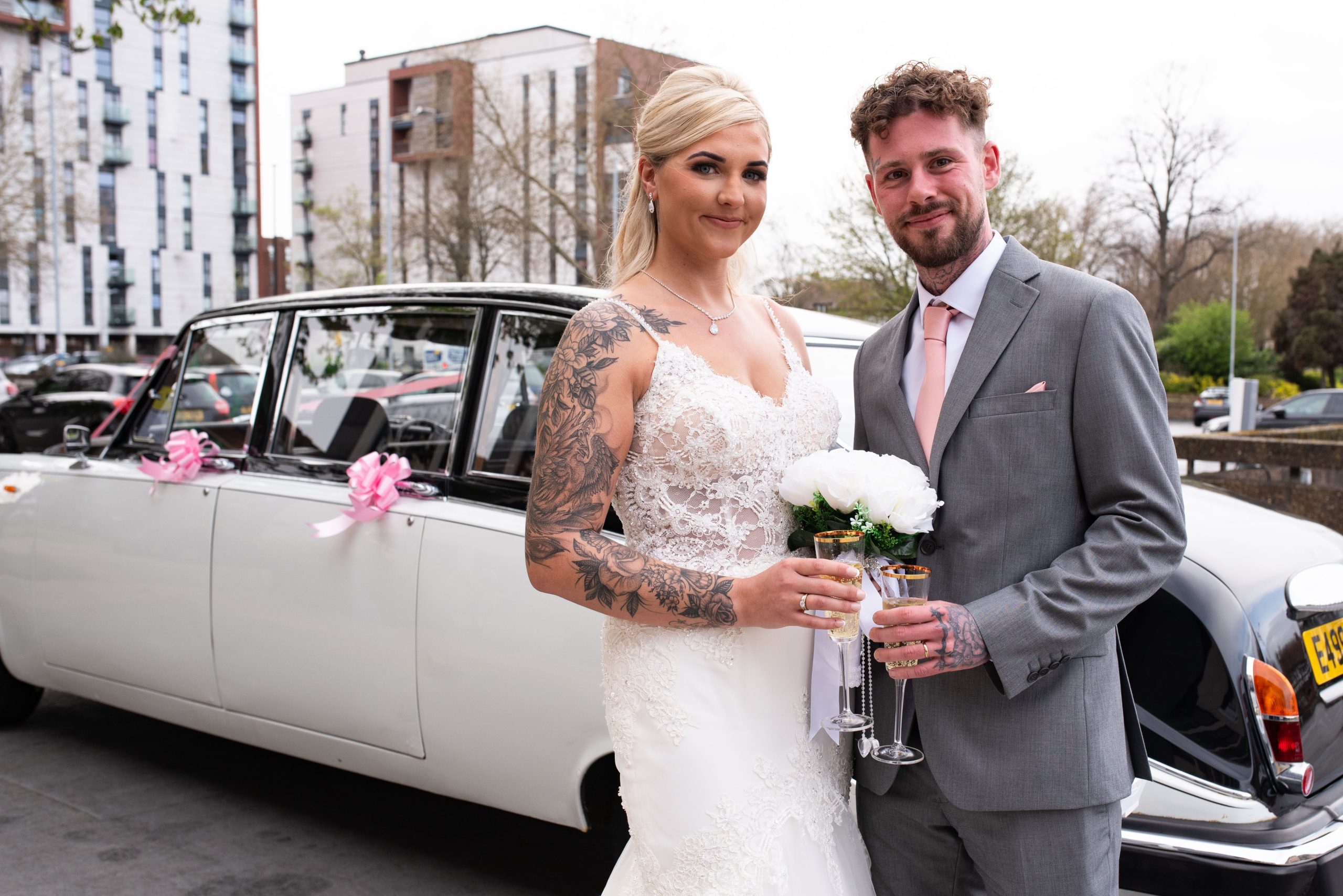 Newlywed couple, bride and groom, holding champagne glasses by a vintage limousine